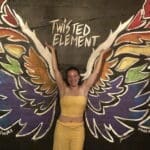 Woman in yellow pants and halter top, standing in front of multi coloured wings and the sign reads "twisted element" and example of dopamine dressing for joy