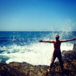 A man wearing pants, standing on big rocks, with the ocean spraying in his face