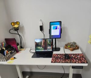 a picture of a small office set up for creativity, with a white sit stand desk, MacBook Air, iPad, mouse pad, and some office supplies and cute knit bee for fun
